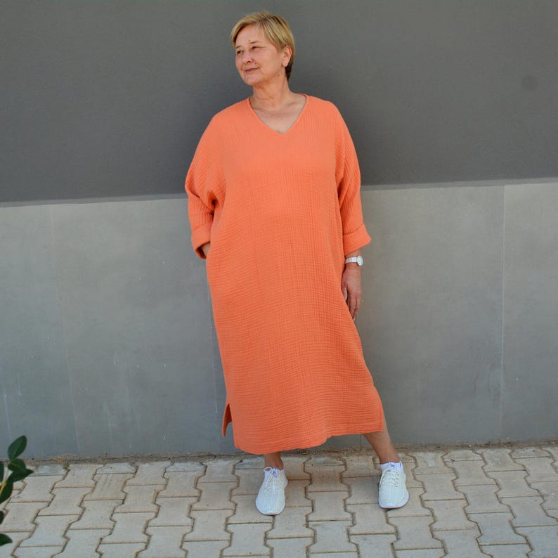 Puede incluir: Un vestido largo de manga larga de color naranja coral con cuello en V y corte holgado. El vestido tiene un aspecto ligeramente fruncido y una abertura lateral. La modelo lleva zapatillas blancas.