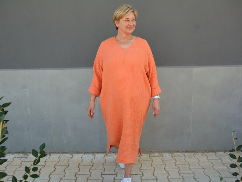 Puede incluir: Una mujer lleva un vestido largo, holgado, de color coral con cuello en V y mangas 3/4. Est&aacute; caminando por un camino de ladrillos frente a una pared gris.