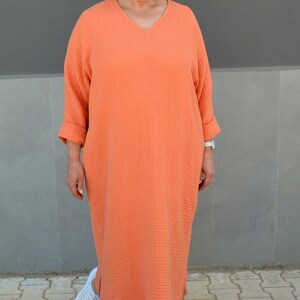 Puede incluir: Una mujer lleva un vestido largo de color naranja coral suelto con cuello en V y mangas 3/4. Lleva zapatillas blancas.