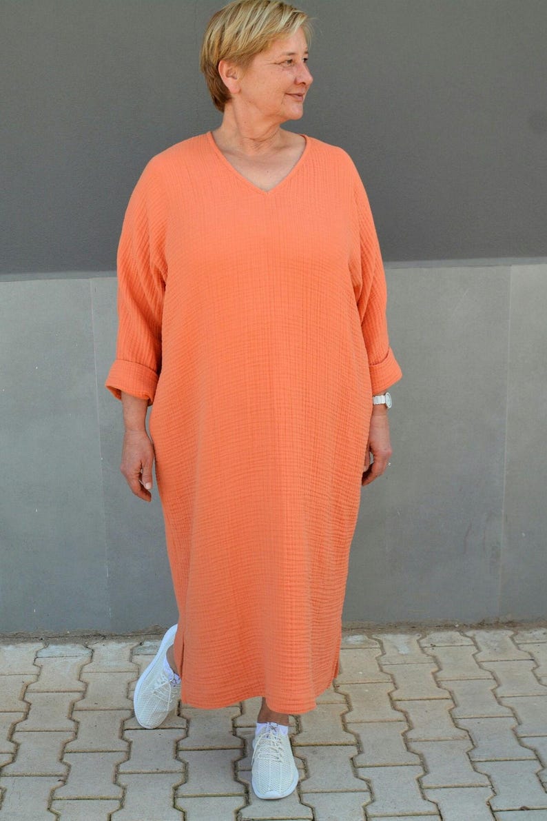 Puede incluir: Una mujer lleva un vestido largo de color naranja coral suelto con cuello en V y mangas 3/4. Lleva zapatillas blancas.