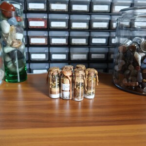 Op de afbeelding: Vijf kleine glazen flesjes met kurkstoppen gevuld met verschillende gedroogde kruiden en bloemen. De flesjes zijn verzegeld met gouden was en zijn in een rij op een houten oppervlak gerangschikt.