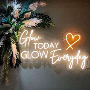 Op de afbeelding: Een wit neonbord met de tekst "Glow Today Glow Everyday" en een hartvorm. Het bord is gemonteerd op een muur met een groene en witte bloemstuk erachter.