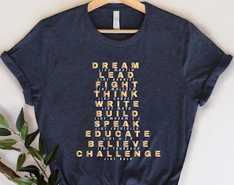 May include: A dark blue t-shirt with a gold print of the words "DREAM LIKE MARTIN", "LEAD LIKE HARRIET", "FIGHT LIKE MALCOLM", "THINK LIKE GARVEY", "WRITE LIKE MAYA", "BUILD LIKE MADAM C", "SPEAK LIKE FREDERICK", "EDUCATE LIKE WEB", "BELIEVE LIKE THURGOOD", and "CHALLENGE LIKE ROSA".