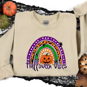 May include: A tan sweatshirt with a rainbow design featuring a pumpkin with a jack-o'-lantern face. The rainbow is made up of different patterns, including leopard print, polka dots, and spiderwebs. The text "Halloween Vibes" is printed below the rainbow.