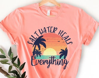 May include: A coral pink t-shirt with a vintage-style graphic of a sunset over the ocean with palm trees. The text "Salt Water Heals Everything" is printed in navy blue.