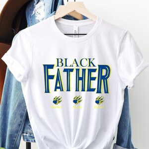 May include: White t-shirt with the text "BLACK FATHER" in blue and yellow lettering. Three paw prints are below the text, each with a name underneath: "Renice", "Aayan", and "Daniel".