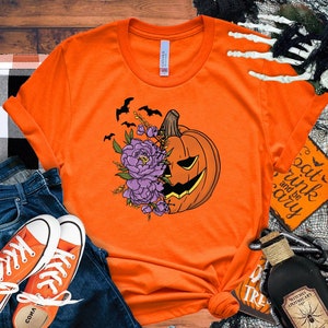 May include: Orange t-shirt with a graphic of a pumpkin with purple flowers and bats. The text "Eat, Drink and be Scary" is on a sign in the background.