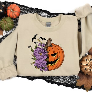 May include: A beige sweatshirt with a graphic of a jack-o'-lantern with purple flowers and bats. The pumpkin is orange with a black face and a white smile.