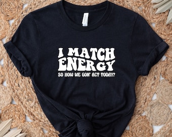 May include: Black t-shirt with white text that reads "I match energy so how we gon' act today?" The text is in a retro font style.
