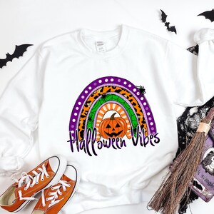 May include: White sweatshirt with a rainbow design featuring a pumpkin, spiders, and the text "Halloween Vibes".