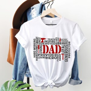 May include: White t-shirt with a red and black word cloud design that spells out "DAD". The words in the cloud include "hard working", "handsome", "strong", "honest", "inspire", "teach", "motivate", "leader", "tough", "patient", "devoted", "brave", "loving", "hero", "provider", "funny", "handy", "loyal", "generous", "protector".