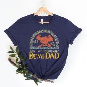 May include: Navy blue t-shirt featuring a graphic of a muscular figure in the ocean, with the text "JUST AN ORDINARY DEMI-DAD" in yellow. The design is framed by an ornate, arched border. The shirt is hanging on a wooden hanger.