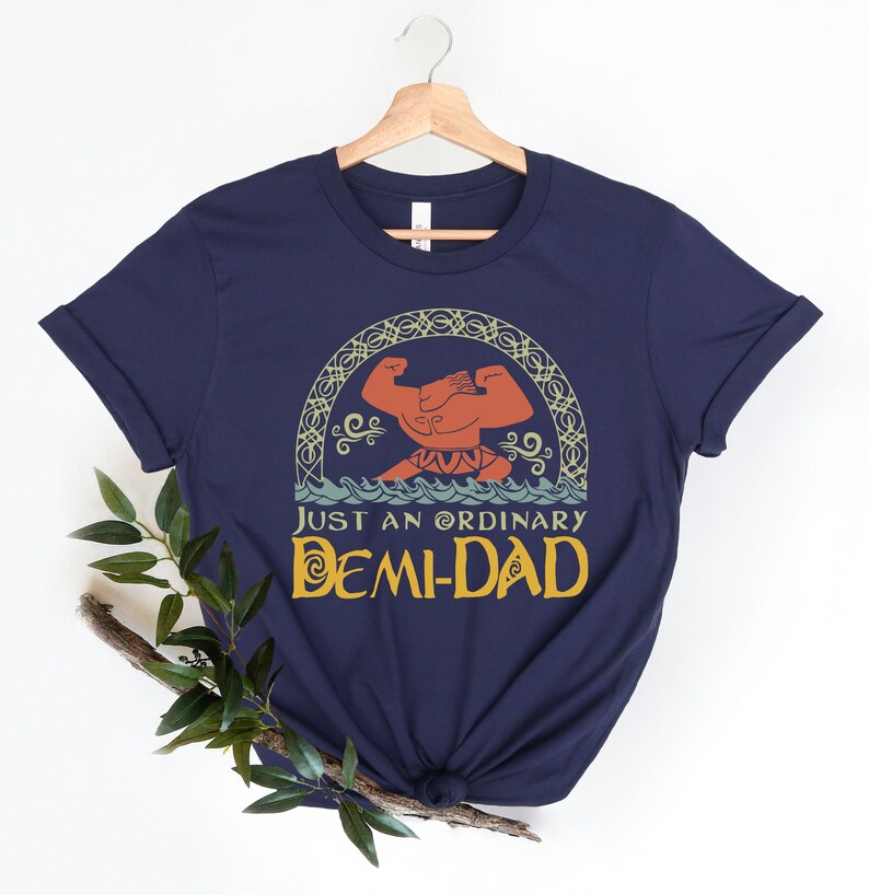 May include: Navy blue t-shirt featuring a graphic of a muscular figure in the ocean, with the text "JUST AN ORDINARY DEMI-DAD" in yellow. The design is framed by an ornate, arched border. The shirt is hanging on a wooden hanger.