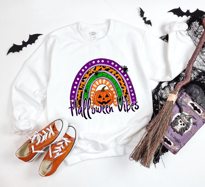 May include: White sweatshirt with a rainbow design featuring a pumpkin, spiders, and the text "Halloween Vibes".