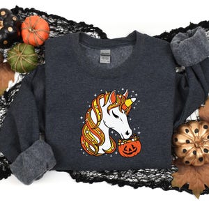 May include: A dark gray sweatshirt with a white unicorn holding a candy corn filled pumpkin. The unicorn has a yellow and orange mane and tail.