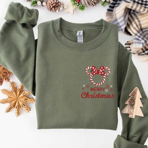 May include: Olive green sweatshirt with a candy cane Mickey Mouse design and the words "Merry Christmas" in red. A wooden Christmas tree is attached to the sleeve. Festive holiday apparel.