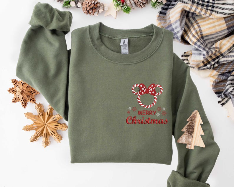 May include: Olive green sweatshirt with a candy cane Mickey Mouse design and the words "Merry Christmas" in red. A wooden Christmas tree is attached to the sleeve. Festive holiday apparel.