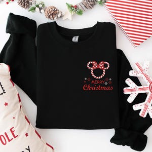 May include: Black crewneck sweatshirt with a red and white Minnie Mouse bow design and the words "Merry Christmas" in red. The sweatshirt is folded on a white surface with Christmas decorations.
