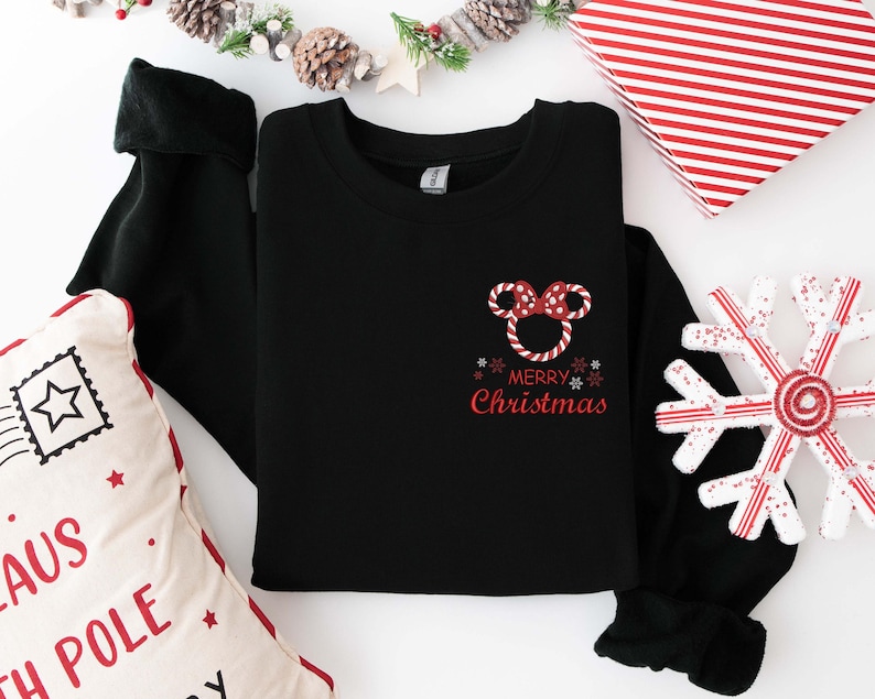 May include: Black crewneck sweatshirt with a red and white Minnie Mouse bow design and the words "Merry Christmas" in red. The sweatshirt is folded on a white surface with Christmas decorations.