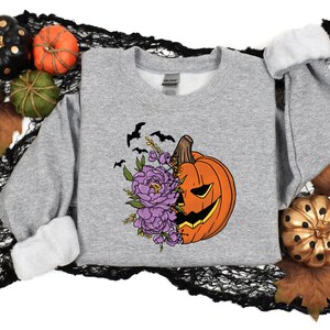 May include: A gray sweatshirt with a graphic of a jack-o'-lantern pumpkin with purple flowers and bats. The pumpkin has a carved face and is partially covered by the flowers.