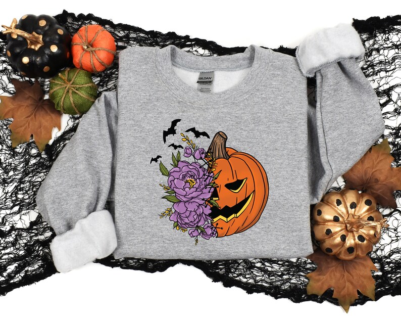 May include: A gray sweatshirt with a graphic of a jack-o'-lantern pumpkin with purple flowers and bats. The pumpkin has a carved face and is partially covered by the flowers.