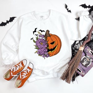 May include: White sweatshirt with a graphic of a jack-o'-lantern with purple flowers. The jack-o'-lantern is smiling and has a carved out face. The flowers are in bloom and have green leaves. There are bats flying around the pumpkin.