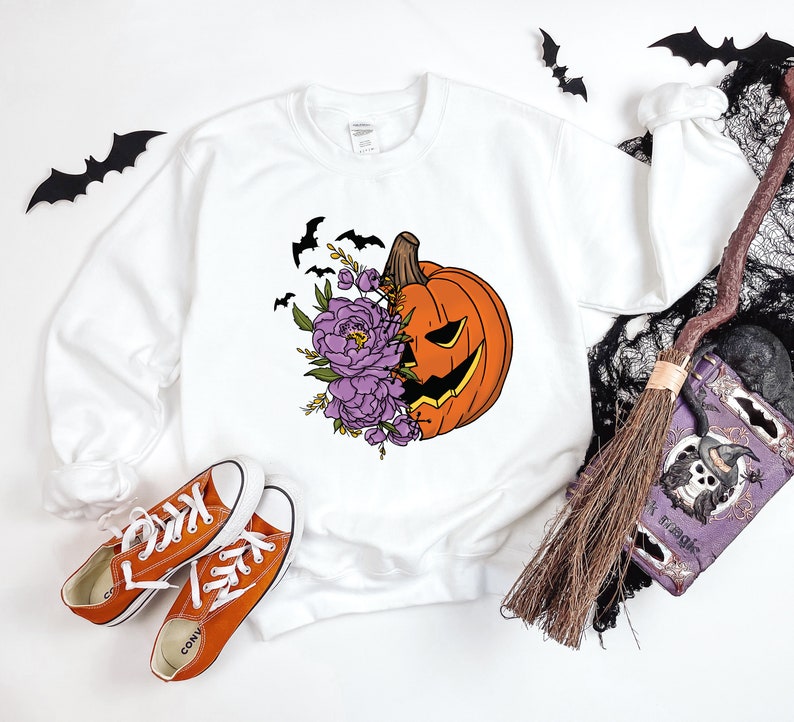 May include: White sweatshirt with a graphic of a jack-o'-lantern with purple flowers. The jack-o'-lantern is smiling and has a carved out face. The flowers are in bloom and have green leaves. There are bats flying around the pumpkin.