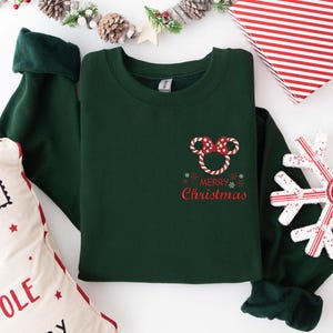 May include: A dark green sweatshirt with a red and white Mickey Mouse-themed design that says "Merry Christmas." The sweatshirt has long sleeves with dark green cuffs. A decorative snowflake and a pillow with text are also in the image.