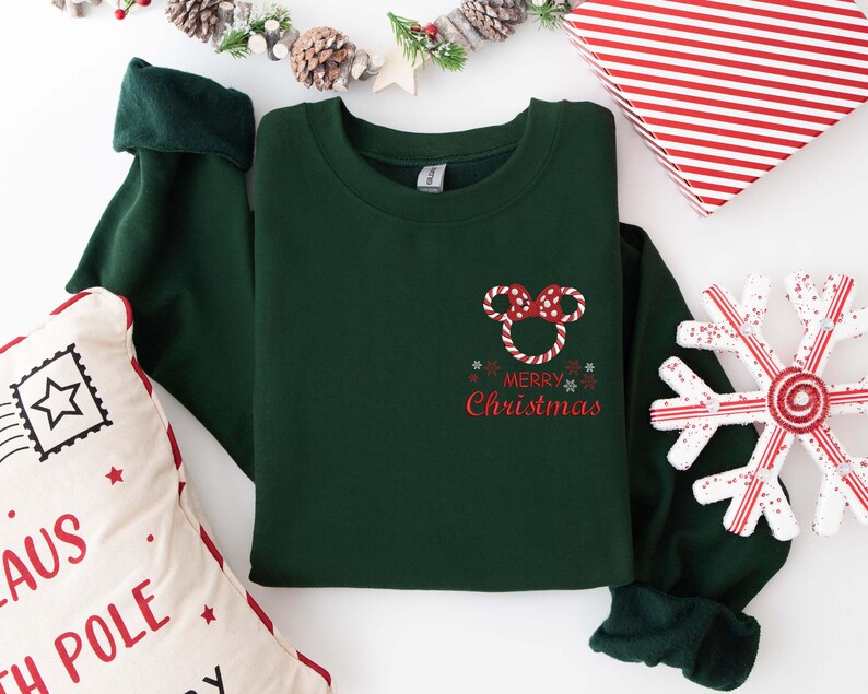 May include: A dark green sweatshirt with a red and white Mickey Mouse-themed design that says "Merry Christmas." The sweatshirt has long sleeves with dark green cuffs. A decorative snowflake and a pillow with text are also in the image.