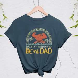 May include: A navy blue t-shirt with the text "JUST AN ORDINARY DEMI-DAD" in yellow and a graphic of a muscular figure. The graphic is framed by a decorative arch. The shirt is hanging on a wooden hanger.