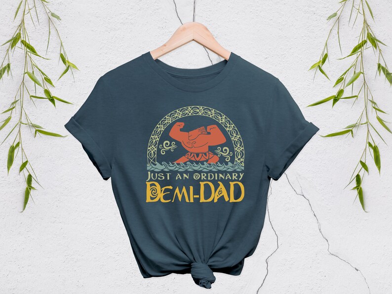 May include: A navy blue t-shirt with the text "JUST AN ORDINARY DEMI-DAD" in yellow and a graphic of a muscular figure. The graphic is framed by a decorative arch. The shirt is hanging on a wooden hanger.