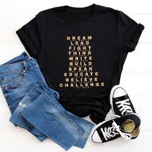 K&ouml;nnte beinhalten: Schwarzes T-Shirt mit goldenem Text, der "DREAM LIKE MARTIN, LEAD LIKE HARRIET, FIGHT LIKE MALCOLM, THINK LIKE THURGOOD, WRITE LIKE MALTA, BUILD LIKE MADAM CJ, SPEAK LIKE FREDERICK, EDUCATE LIKE W.E.B, BELIEVE LIKE THURGOOD, CHALLENGE LIKE ROJA" lautet.