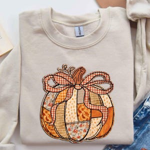 May include: Cream-colored sweatshirt featuring a patchwork pumpkin design. The pumpkin has a brown stem and a bow made of orange and brown plaid fabric. The pumpkin's body is made of various floral and patterned fabrics.
