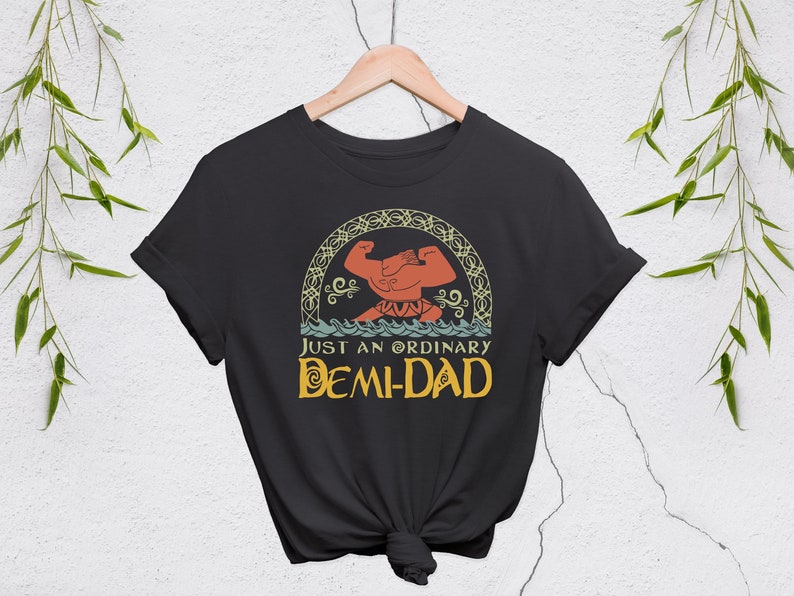 May include: Black t-shirt with a graphic of a muscular figure flexing, set within a decorative arch. The text "JUST AN ORDINARY DEMI-DAD" is below the image, in a stylized font. The shirt is hanging on a wooden hanger.