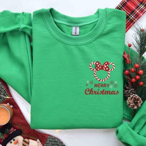 May include: A green crewneck sweatshirt with a candy cane Mickey Mouse design and the words "Merry Christmas" in red. The sweatshirt is folded, with a Christmas stocking and decorations in the background.