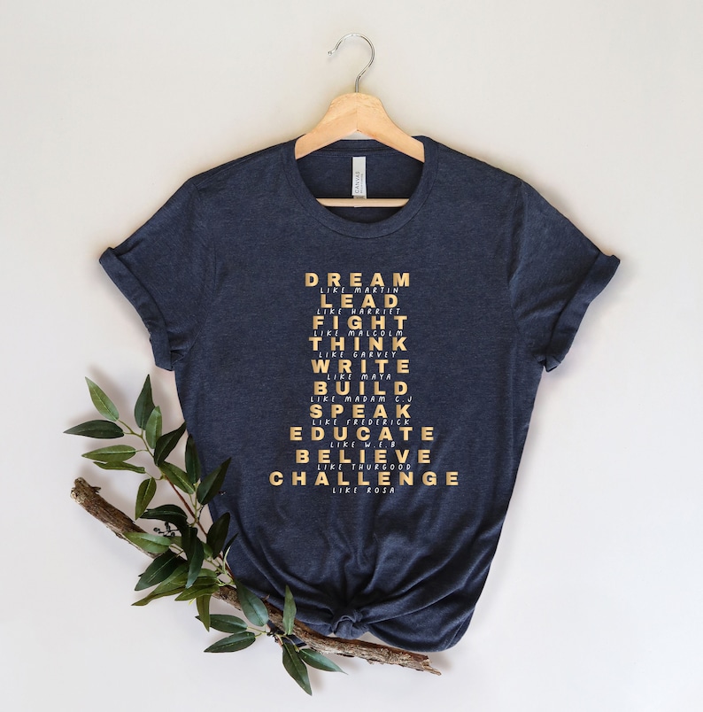 K&ouml;nnte beinhalten: Ein dunkelblaues T-Shirt mit einem goldenen Aufdruck der W&ouml;rter "DREAM LIKE MARTIN", "LEAD LIKE HARRIET", "FIGHT LIKE MALCOLM", "THINK LIKE GARVEY", "WRITE LIKE MAYA", "BUILD LIKE MADAM C", "SPEAK LIKE FREDERICK", "EDUCATE LIKE WEB", "BELIEVE LIKE THURGOOD" und "CHALLENGE LIKE ROSA".