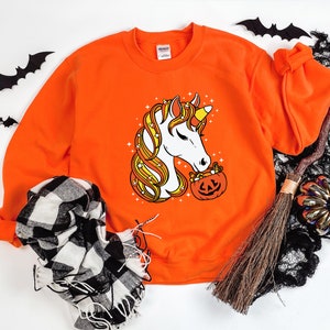 May include: Orange sweatshirt with a white unicorn graphic holding a pumpkin filled with candy corn. The unicorn has a yellow horn and a pink nose.