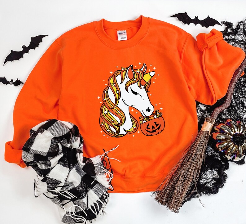 May include: Orange sweatshirt with a white unicorn graphic holding a pumpkin filled with candy corn. The unicorn has a yellow horn and a pink nose.
