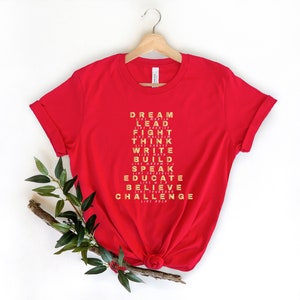 K&ouml;nnte beinhalten: Rotes T-Shirt mit goldfarbenem Text, der "DREAM LIKE MARTIN, LEAD LIKE HARRIET, FIGHT LIKE MALCOLM, THINK LIKE GARVEY, WRITE LIKE MAYA, BUILD LIKE MADAM C.J., SPEAK LIKE FREDERICK, EDUCATE LIKE W.E.B., BELIEVE LIKE THURGOOD, CHALLENGE LIKE ROSA" lautet.