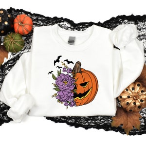 May include: White sweatshirt with a graphic of a pumpkin with purple flowers and bats. The pumpkin has a scary face.
