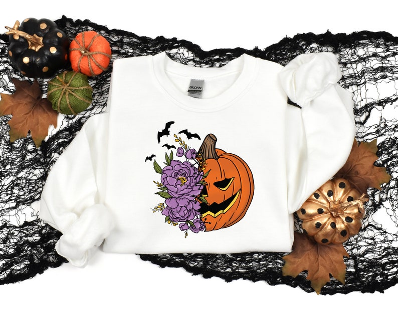 May include: White sweatshirt with a graphic of a pumpkin with purple flowers and bats. The pumpkin has a scary face.