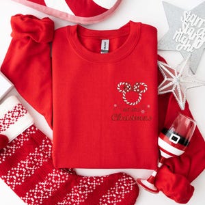 May include: A red crewneck sweatshirt with a candy cane Mickey Mouse design and the words "Merry Christmas." The image includes a Christmas stocking, a Santa hat, and a decorative star.