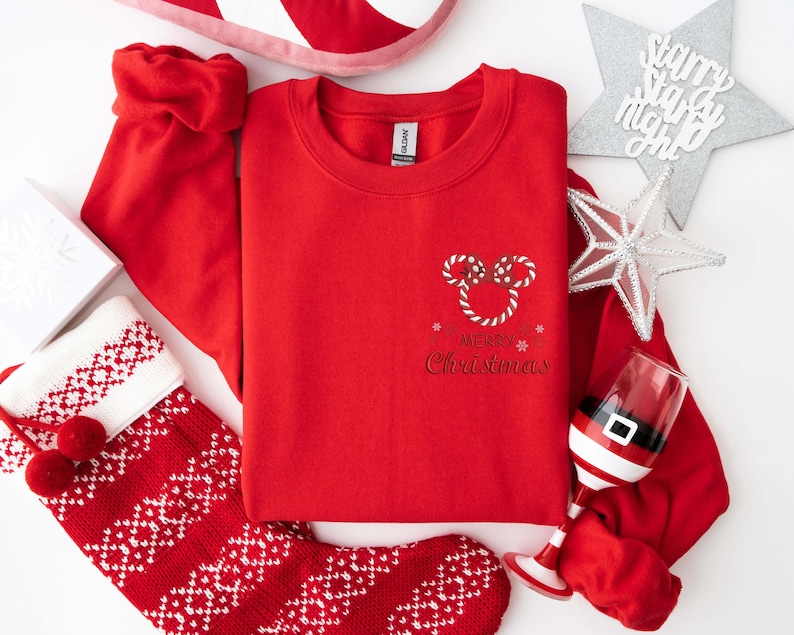 May include: A red crewneck sweatshirt with a candy cane Mickey Mouse design and the words "Merry Christmas." The image includes a Christmas stocking, a Santa hat, and a decorative star.