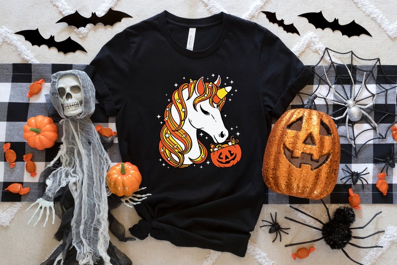 May include: Black t-shirt with a white unicorn graphic. The unicorn is made of candy corn and is holding a jack-o'-lantern filled with candy corn. The shirt is surrounded by Halloween decorations, including a spider web, a ghost, and candy corn.