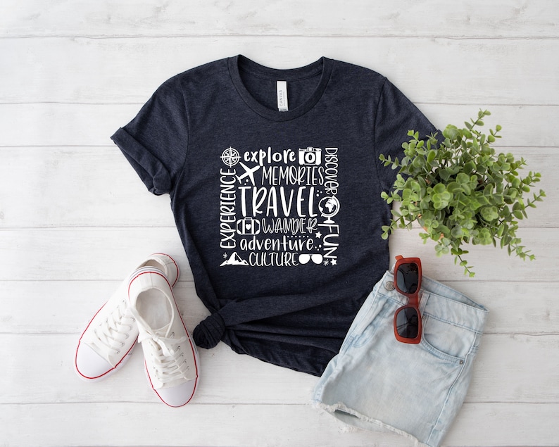 May include: A navy blue t-shirt with white text that reads "Experience, Explore, Memories, Travel, Wander, Discover, Adventure, Culture, Fun". The text is arranged in a vertical column with a compass, airplane, camera, and globe icons.