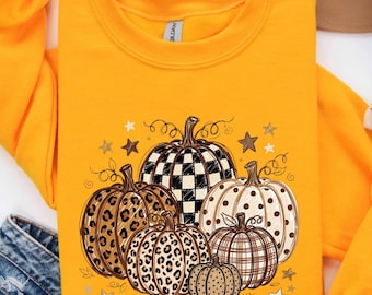 Könnte beinhalten: Ein leuchtend orangefarbenes Sweatshirt mit Rundhalsausschnitt und einem Grafikdruck von fünf Kürbissen. Die Kürbisse zeigen verschiedene Muster wie schwarz-weißes Schachbrett, Leopardenmuster, Punkte und Karos, umgeben von kleinen Sternen.