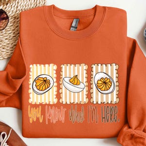 May include: Orange sweatshirt with a graphic of three deviled eggs on striped backgrounds. The text "YOU KNOW WHY I'M HERE" is printed in a yellow-orange color below the eggs. The sweatshirt is a solid orange color.