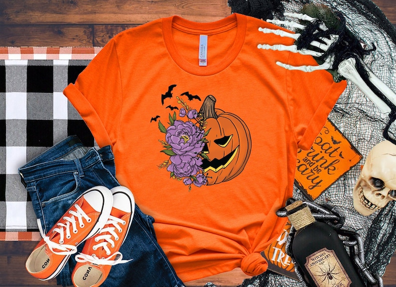 May include: Orange t-shirt with a graphic of a pumpkin with purple flowers and bats. The text "Eat, Drink and be Scary" is on a sign in the background.