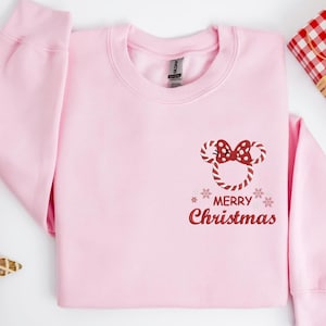 May include: A light pink sweatshirt with a crew neck. The front features a candy cane Mickey Mouse design with a red bow and the words "MERRY Christmas" in red script. Two snowflakes are on either side of the text.