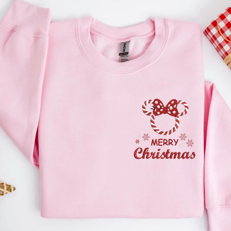 May include: A light pink sweatshirt with a crew neck. The front features a candy cane Mickey Mouse design with a red bow and the words "MERRY Christmas" in red script. Two snowflakes are on either side of the text.